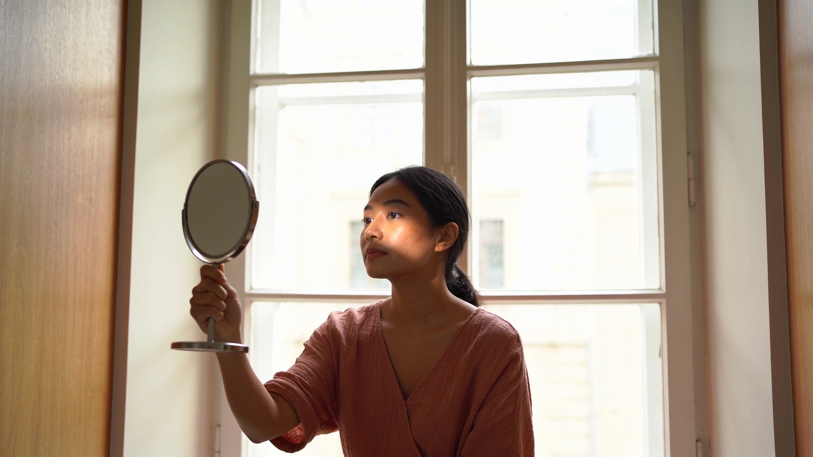 A Southeast Asian woman in her late twenties checking her cheek in a hand mirror by a window in soft side light, her whole head in frame