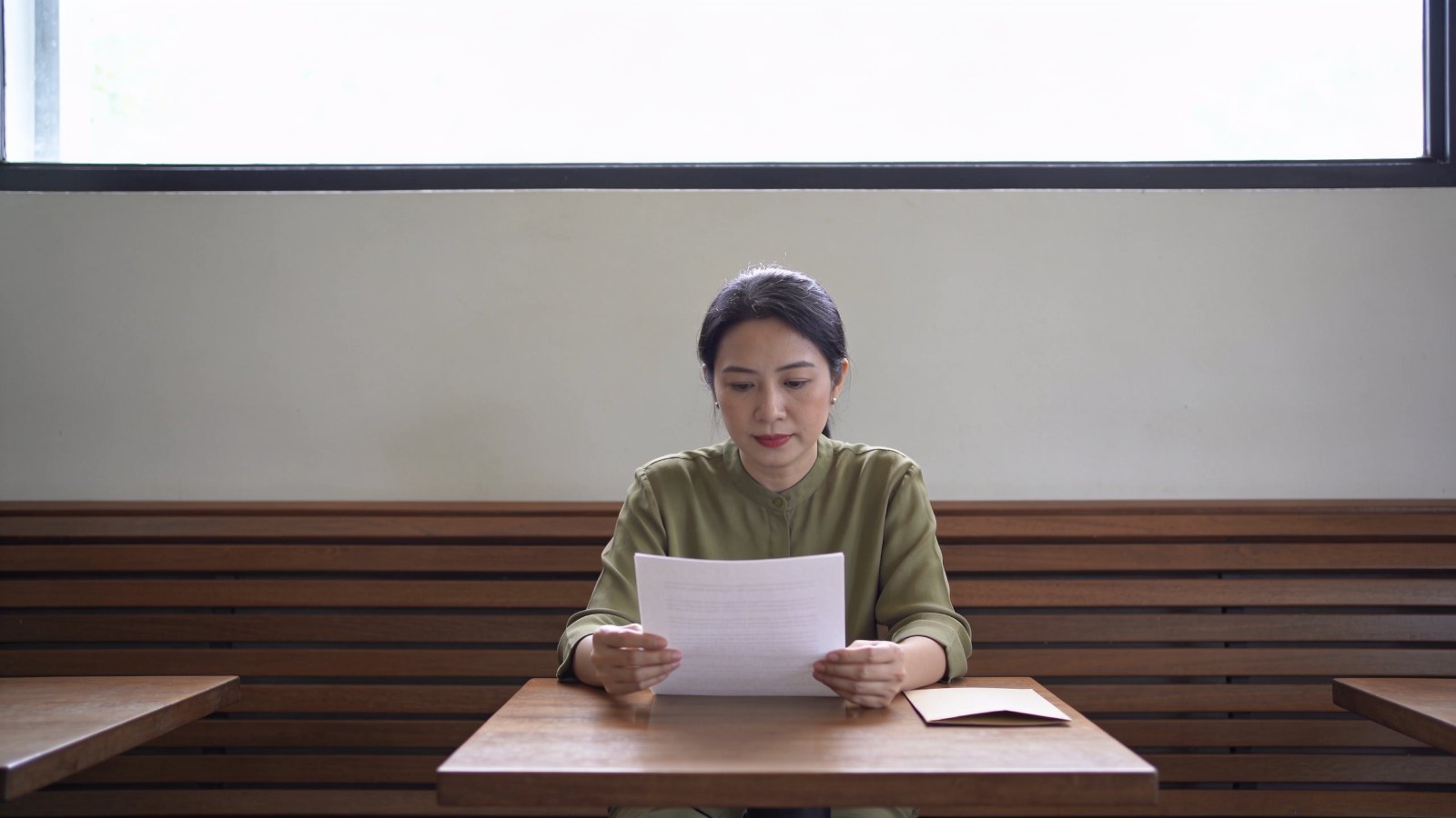 A Chinese Malaysian woman in her thirties reading a printed treatment record at a café table, her whole face and head in frame
