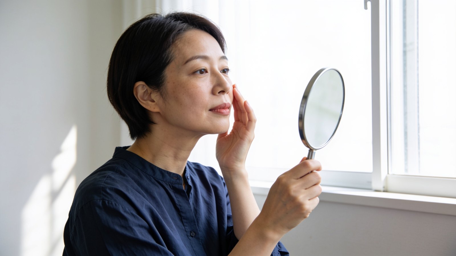 A Japanese woman in her forties living in Singapore looking closely at her cheek in a hand mirror by a window, her whole face and head in frame