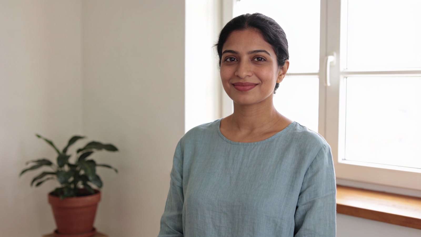 A woman in her thirties with fresh, even skin smiling softly beside a bright window, her whole head and face in frame