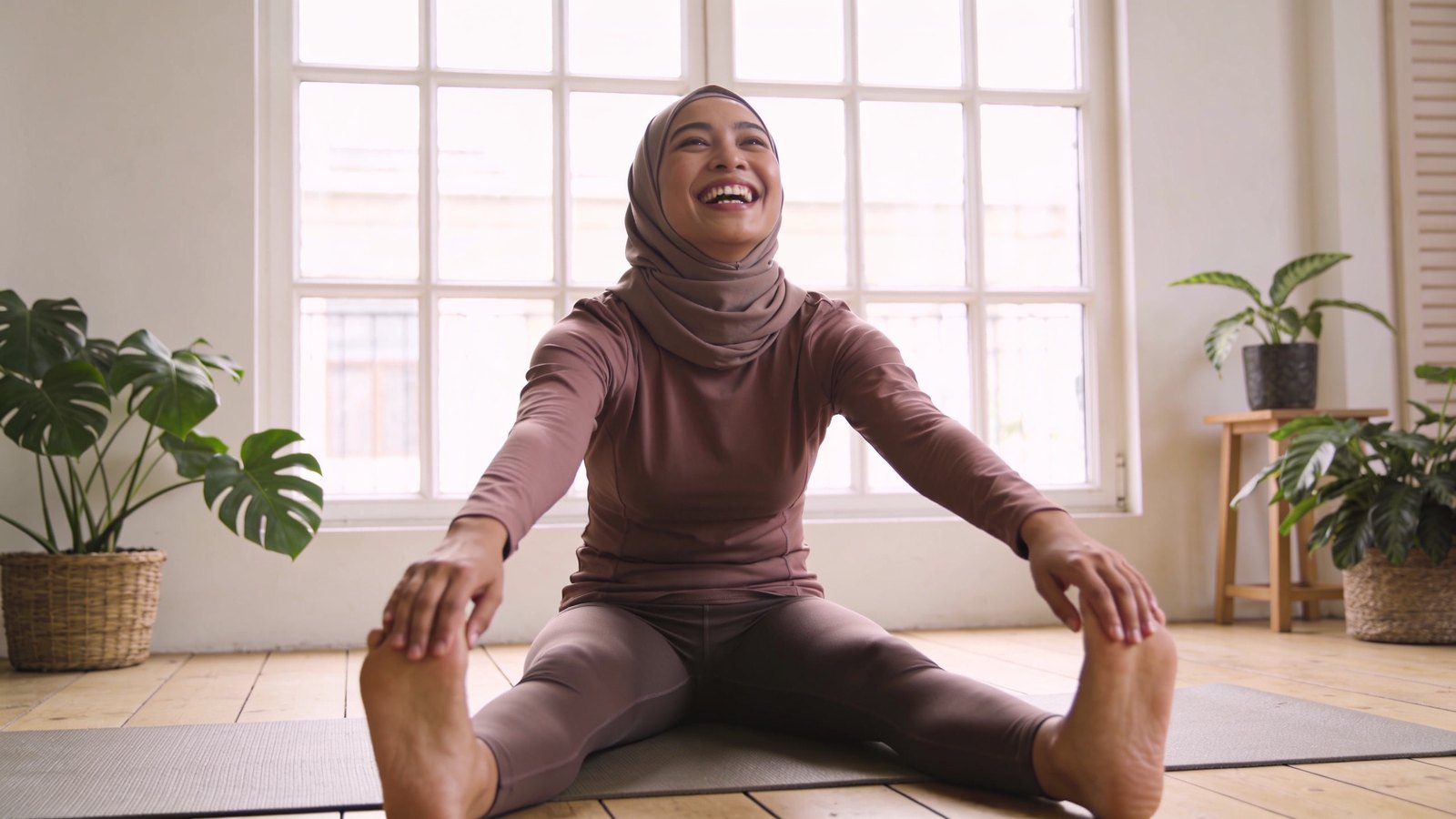 A Southeast Asian woman in her thirties in modest activewear laughing during a stretching class in a bright studio, her whole head and face in frame