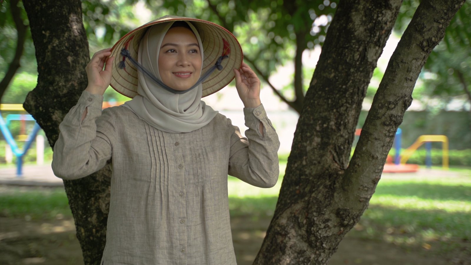 A Singaporean mother in her thirties putting a sun hat on her own head beside a sunny playground, her whole face and head in frame