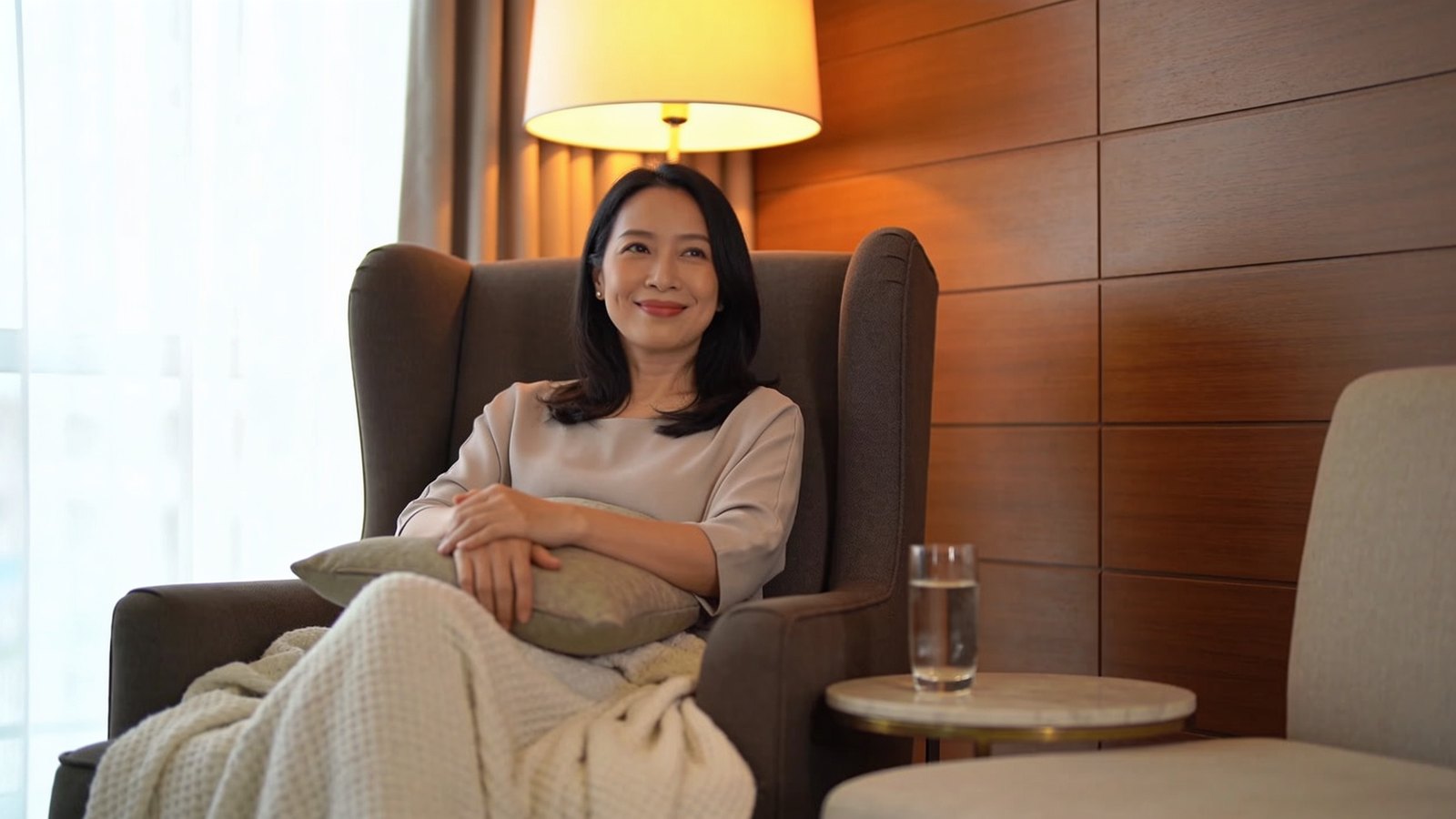 A Chinese Singaporean woman in her thirties resting calmly in a softly lit treatment lounge with a blanket over her lap, her whole face and head in frame