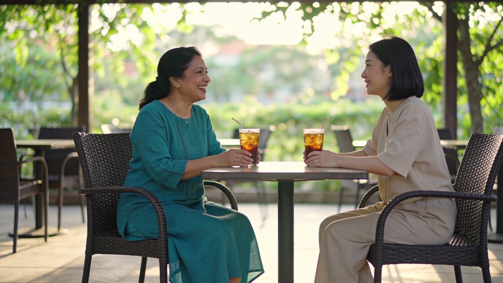 An Indian Singaporean woman in her forties smiling while talking with a friend at an outdoor café table in soft shade, both faces fully in frame