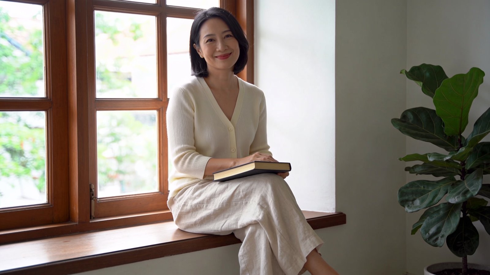 A Chinese Singaporean woman in her forties smiling softly while relaxing at home with a book, her whole face and head in frame