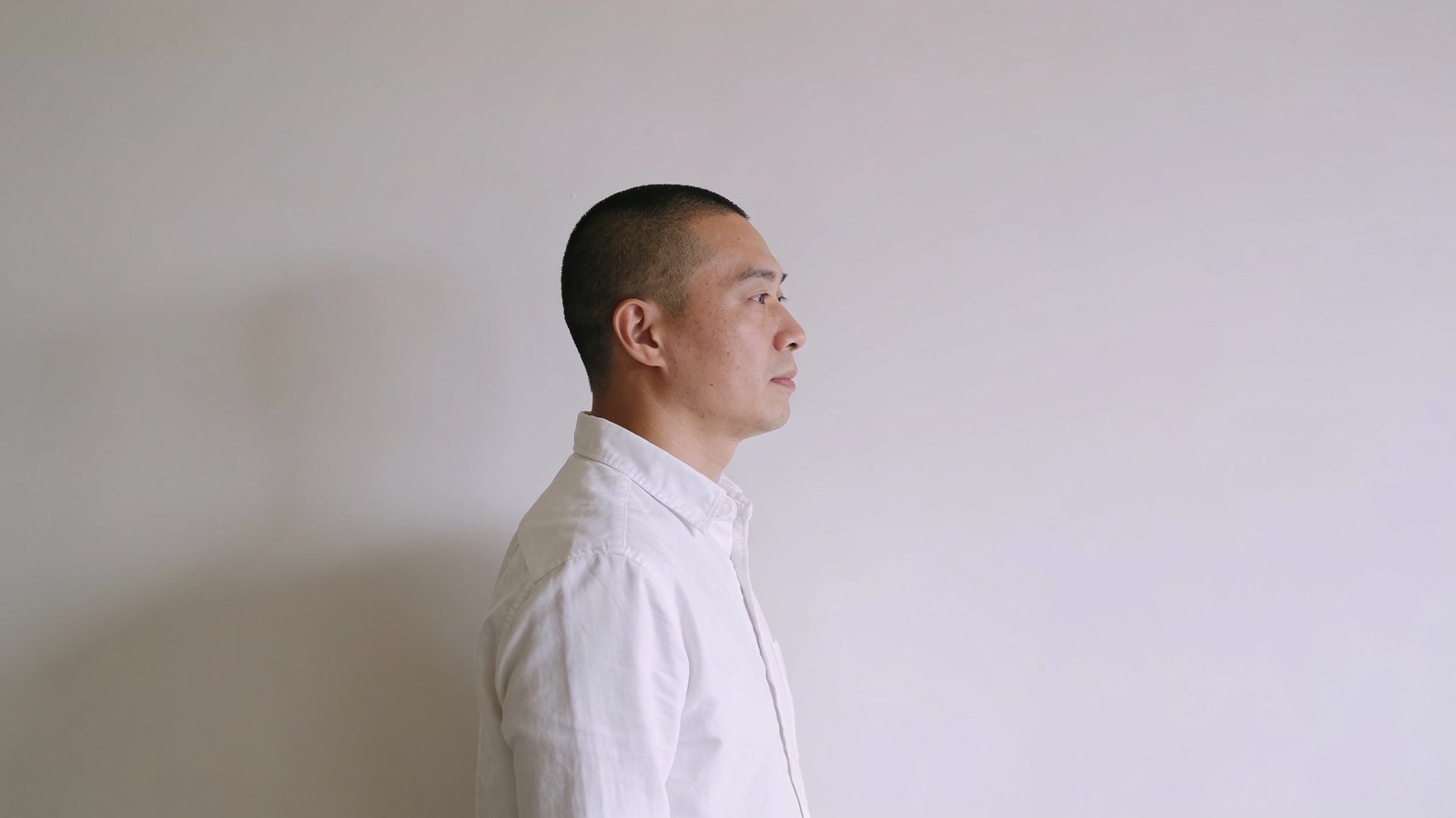 East Asian man in his late thirties with a neat buzz-cut look, seen in profile against a plain light wall, his whole head inside the frame