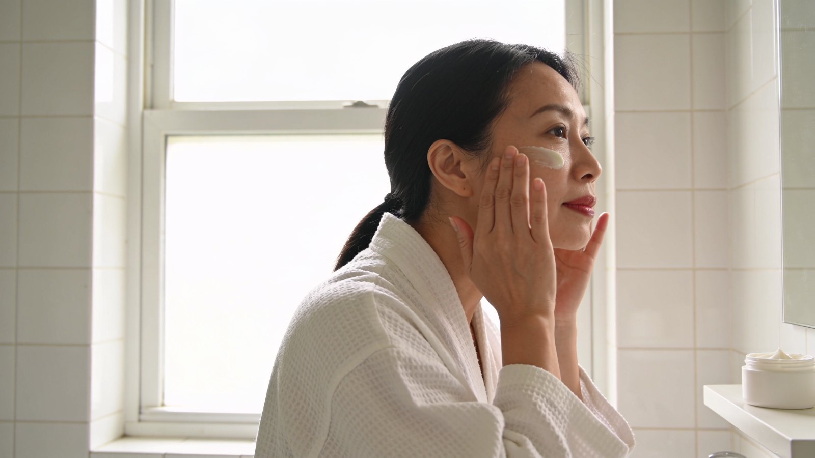 A Chinese Singaporean woman in her forties applying moisturiser at a bright bathroom mirror, her whole face and head in frame