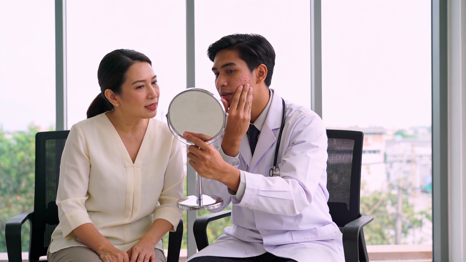 A doctor holding a hand mirror so a patient can see her cheek during an acne scar consultation